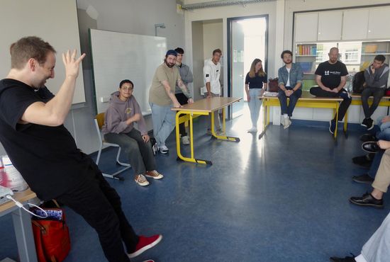 Mehrere Jugendliche in einem Klassenzimmer, sitzend und stehend in einem Kreis. Ein Mann erklärt etwas.